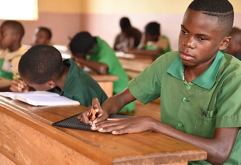 Boy reading braille as the other students in class write notes | © CBM