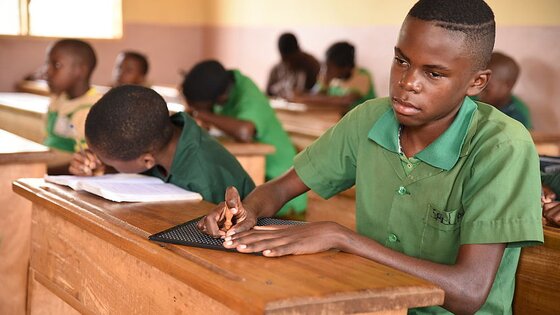 Boy reading braille as the other students in class write notes | © CBM